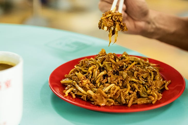 A plate of Char Kway Teow on a blue table with a person's hand using chopsticks.